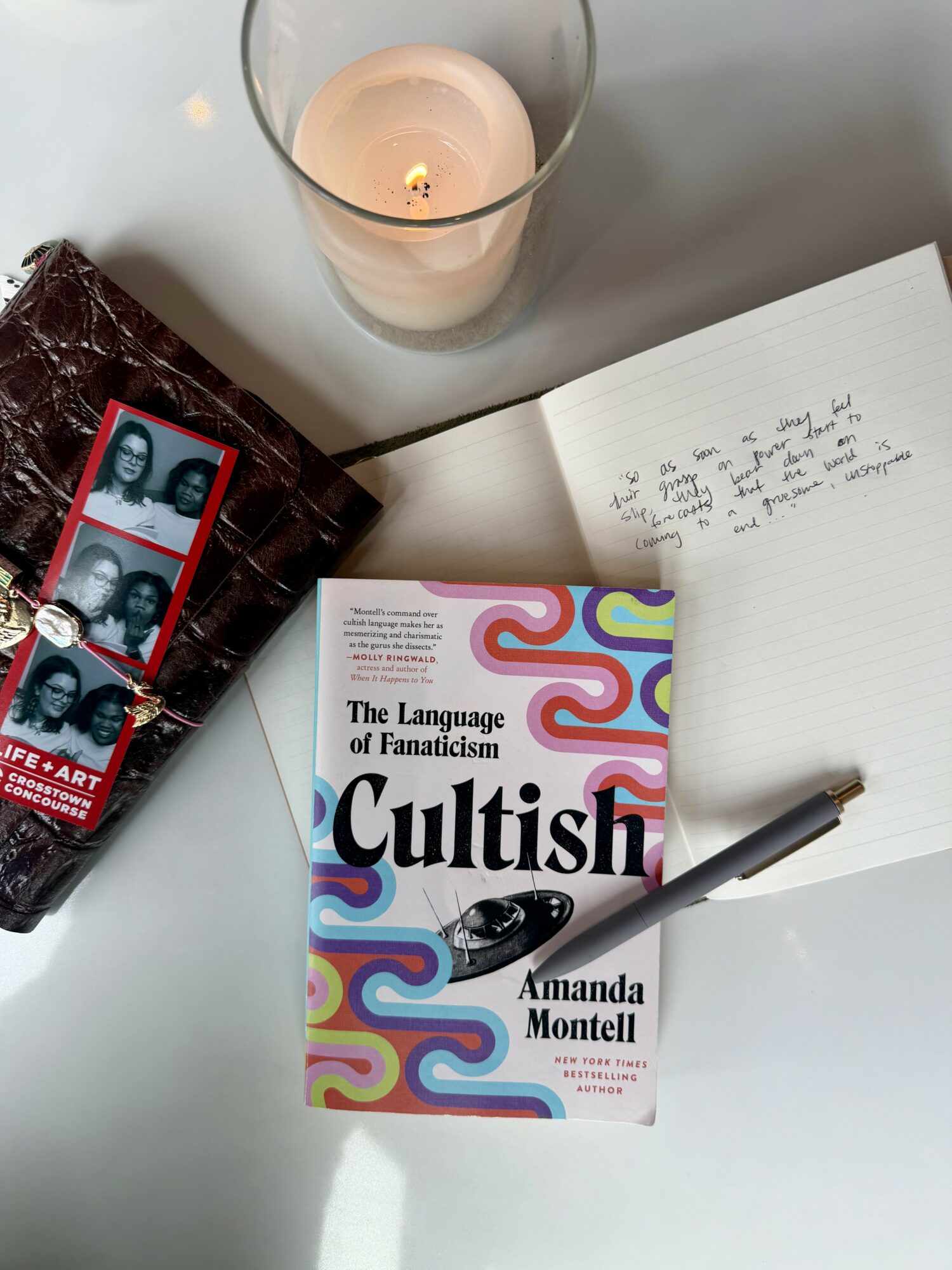 Open notebook with handwritten notes, book titled 'Cultish' by Amanda Montell, photo booth strip, candle, and wallet on table.