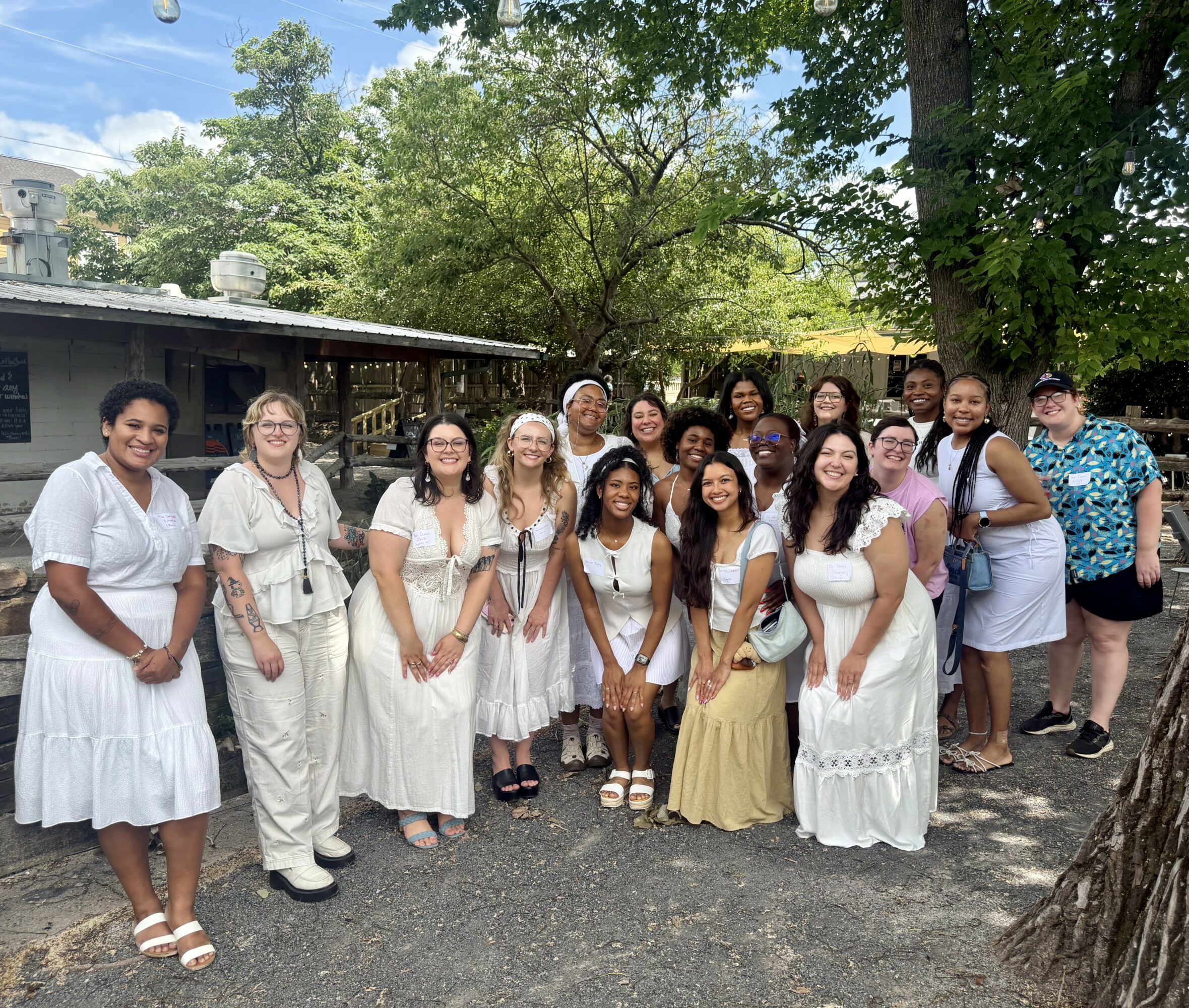 Group of women and children outdoors, some wearing white dresses, standing on a gravel path with trees and a building in background.