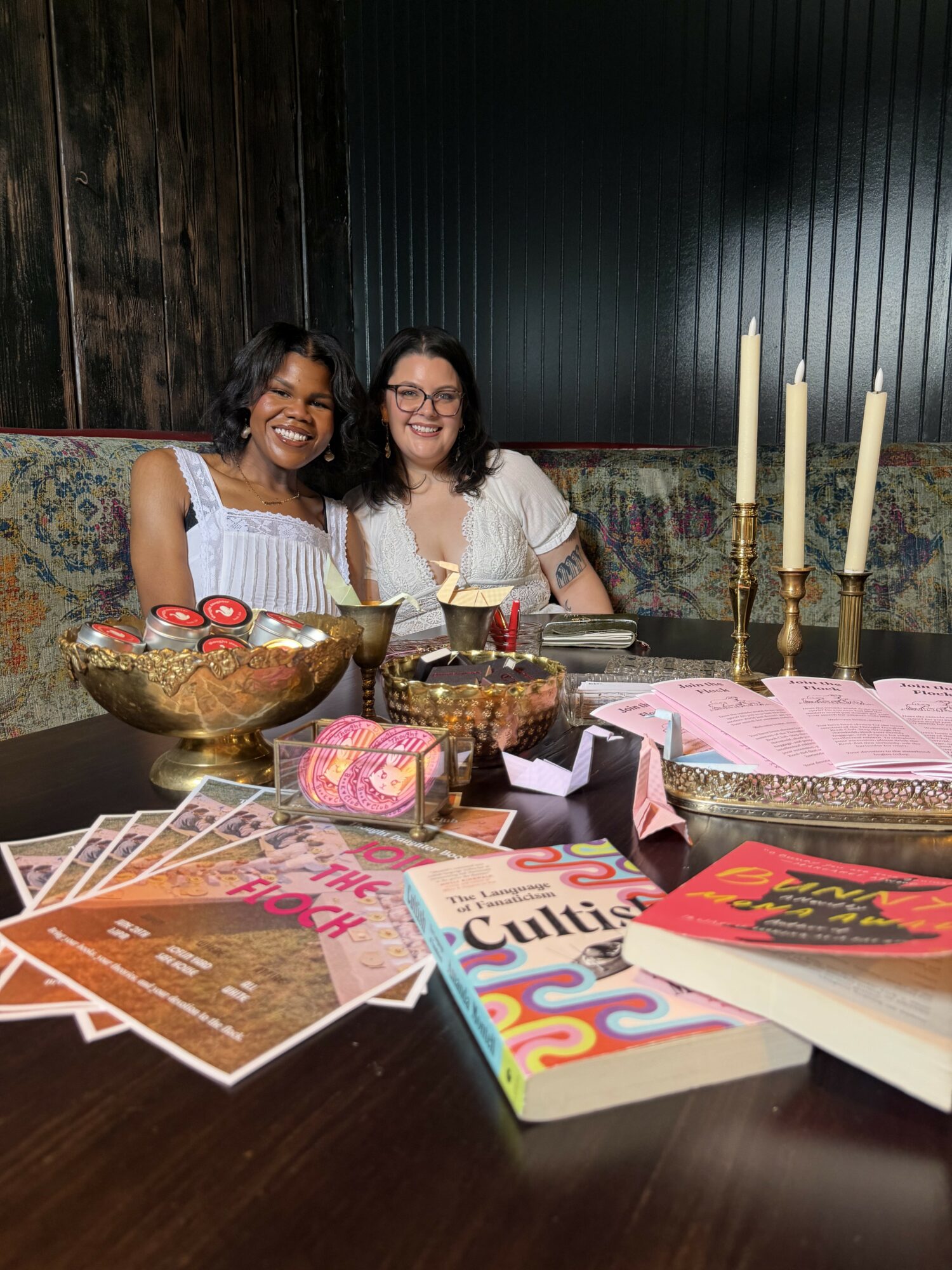 Two women sit at a table with books, candles, and decorative items, smiling at the camera.