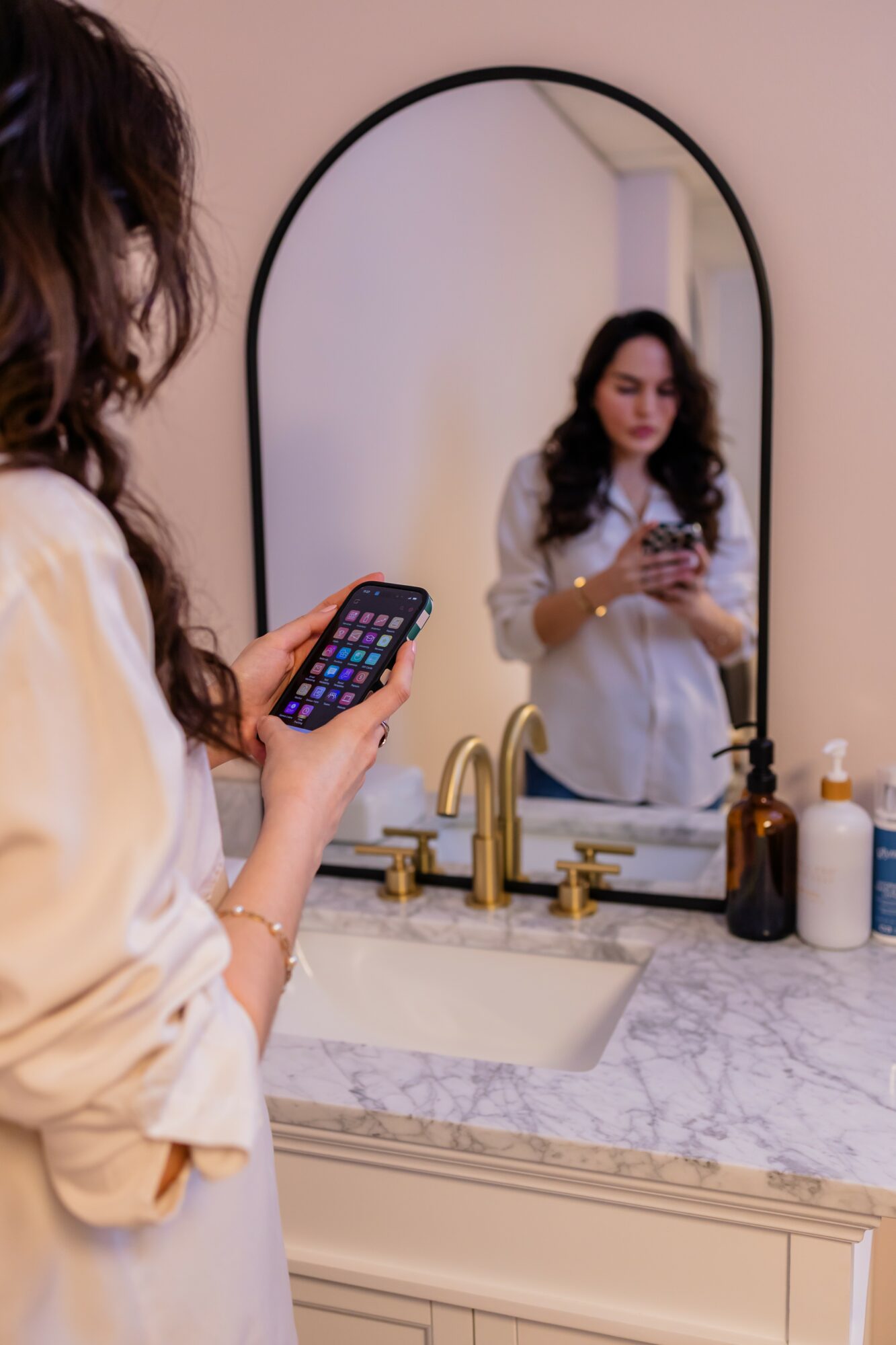Woman with curly hair looking at her phone in front of a mirror above a bathroom sink, with toiletries on the counter.