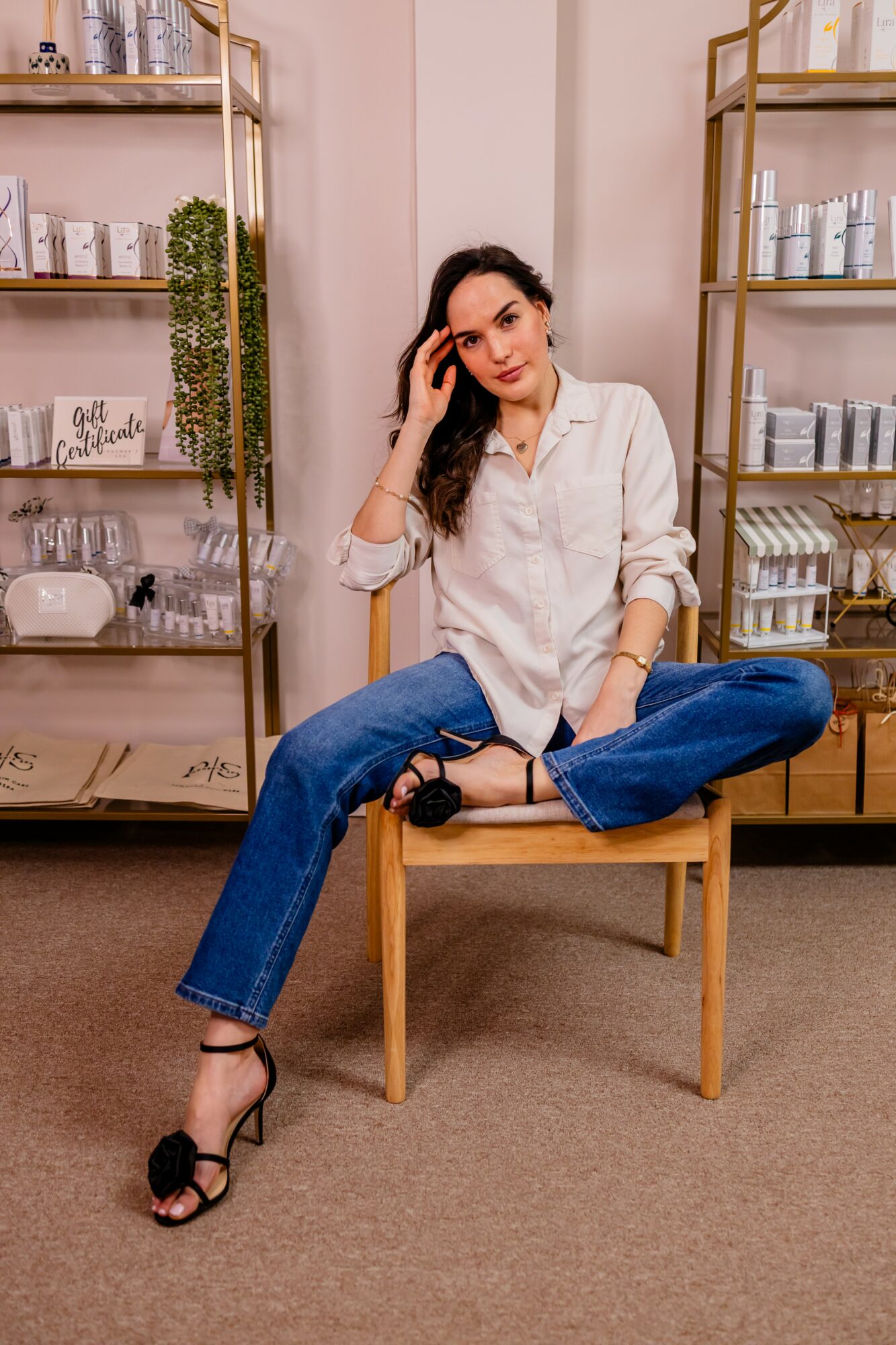 Woman sitting on a chair with legs crossed, in a room with shelves of skincare products, wearing a white shirt and blue jeans.