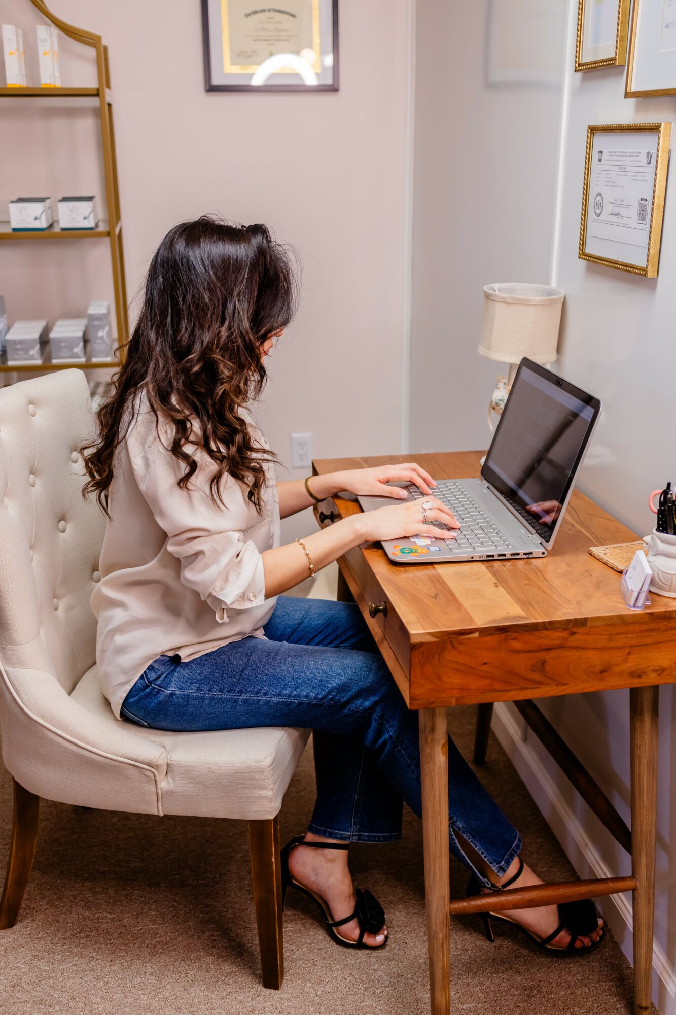 Woman with long dark hair working on a laptop at a wooden desk in a room with framed certificates on the wall.