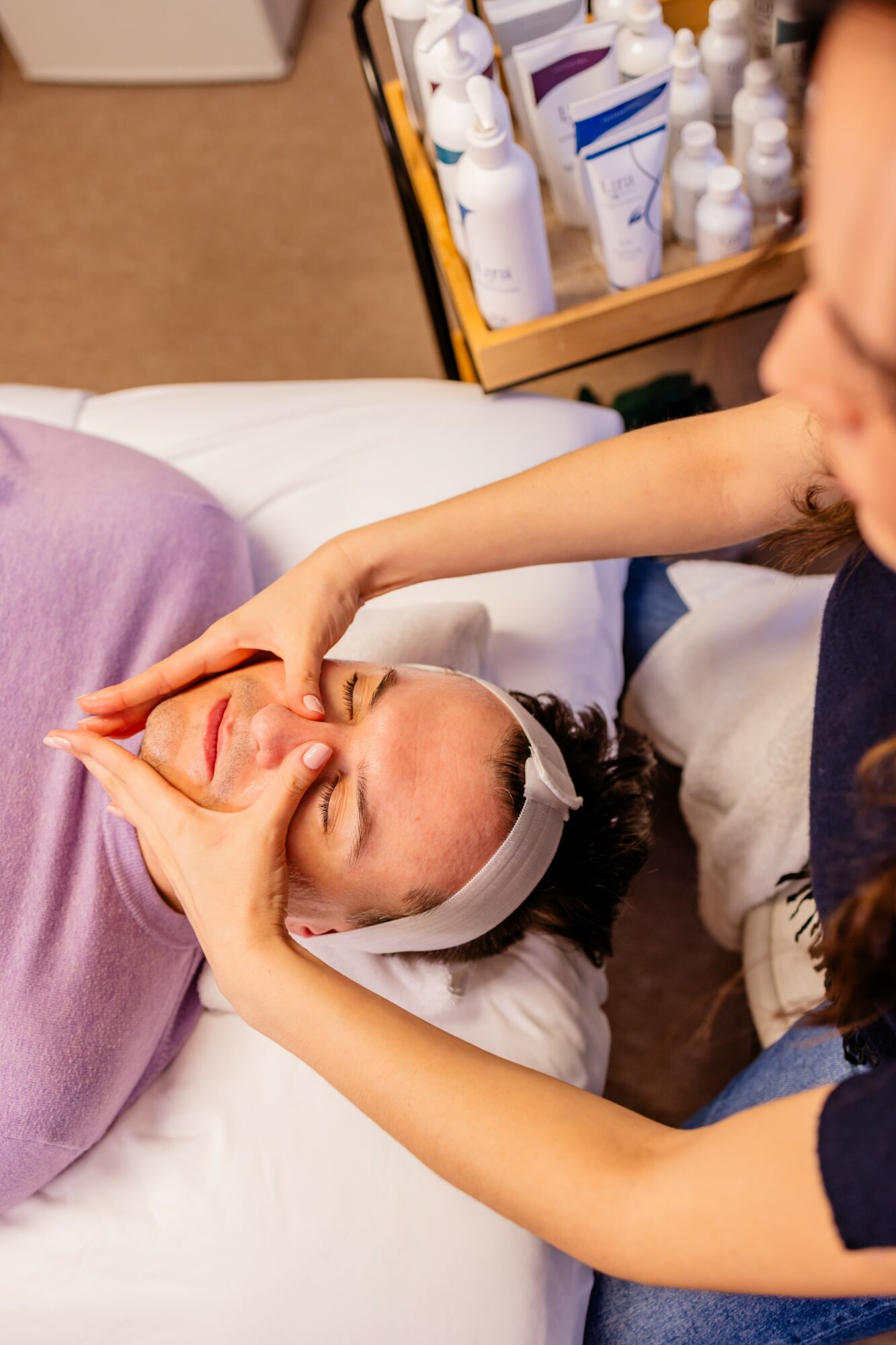 Person receiving facial treatment with eyes closed, lying on a bed, with a practitioner applying a mask or cream, skincare products nearby.
