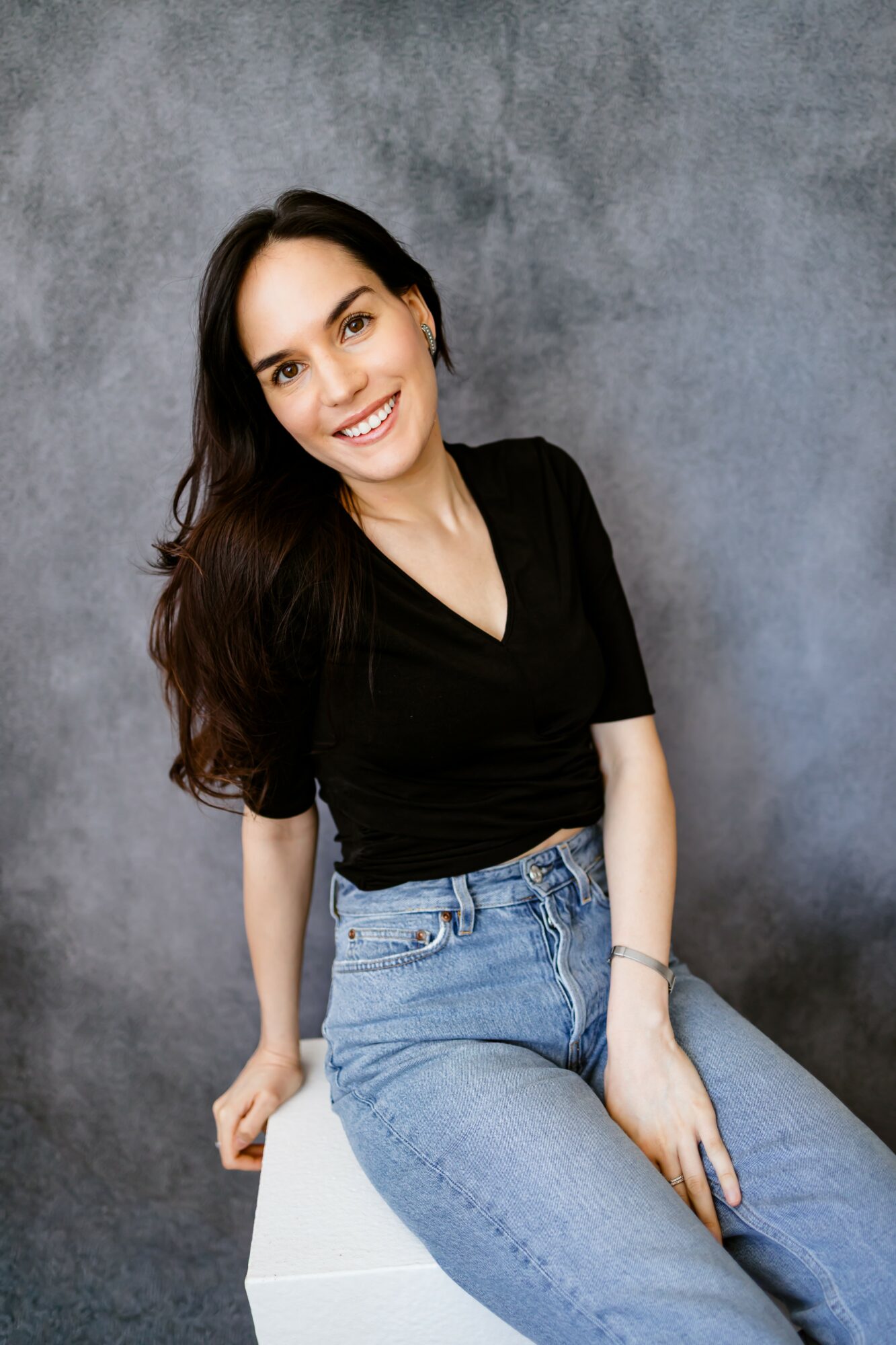 Young woman with long dark hair smiling, wearing a black top and blue jeans, sitting on a white block against a gray background.