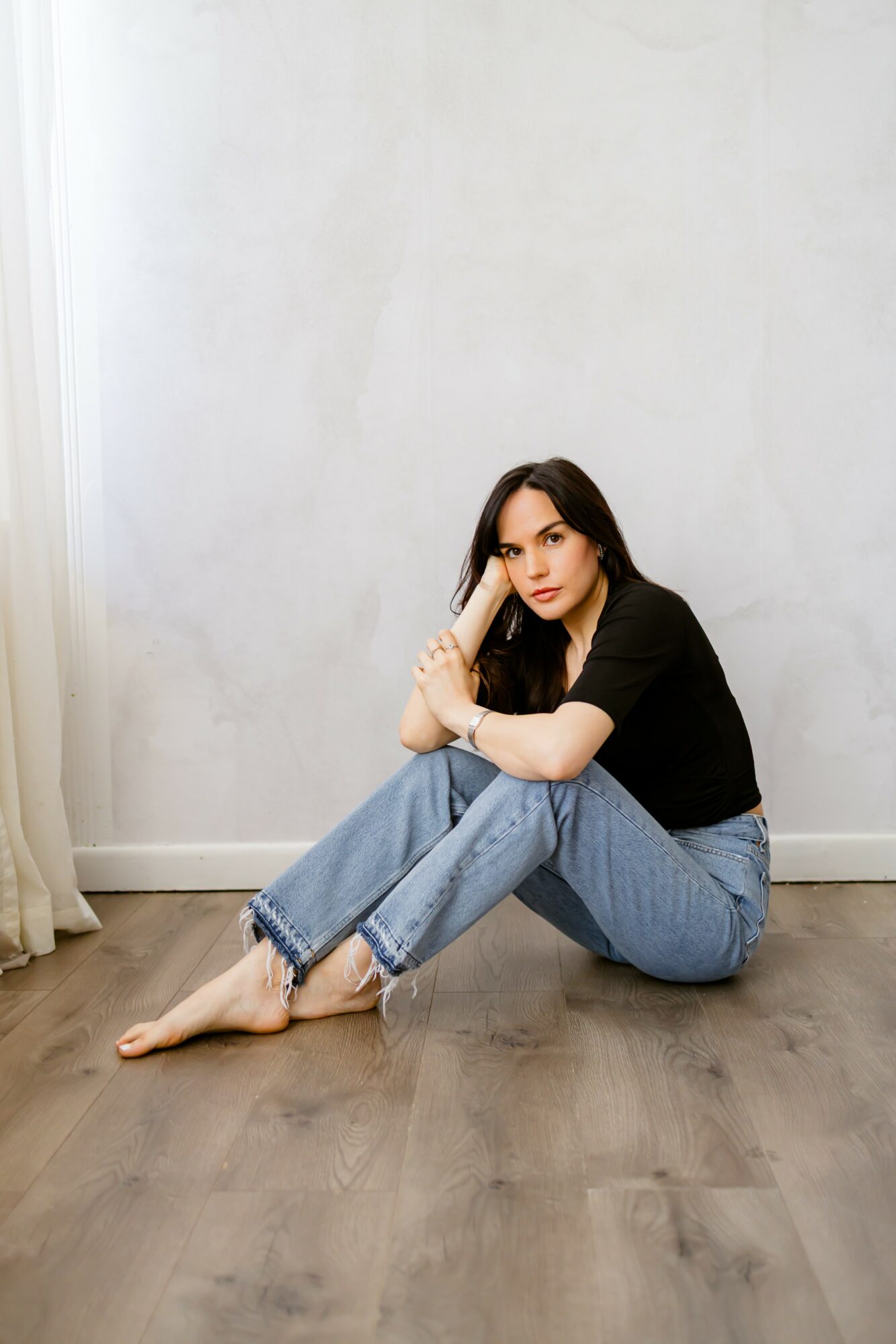 Young woman with dark hair sitting on wooden floor, leaning against a plain white wall, wearing a black shirt and jeans, barefoot.