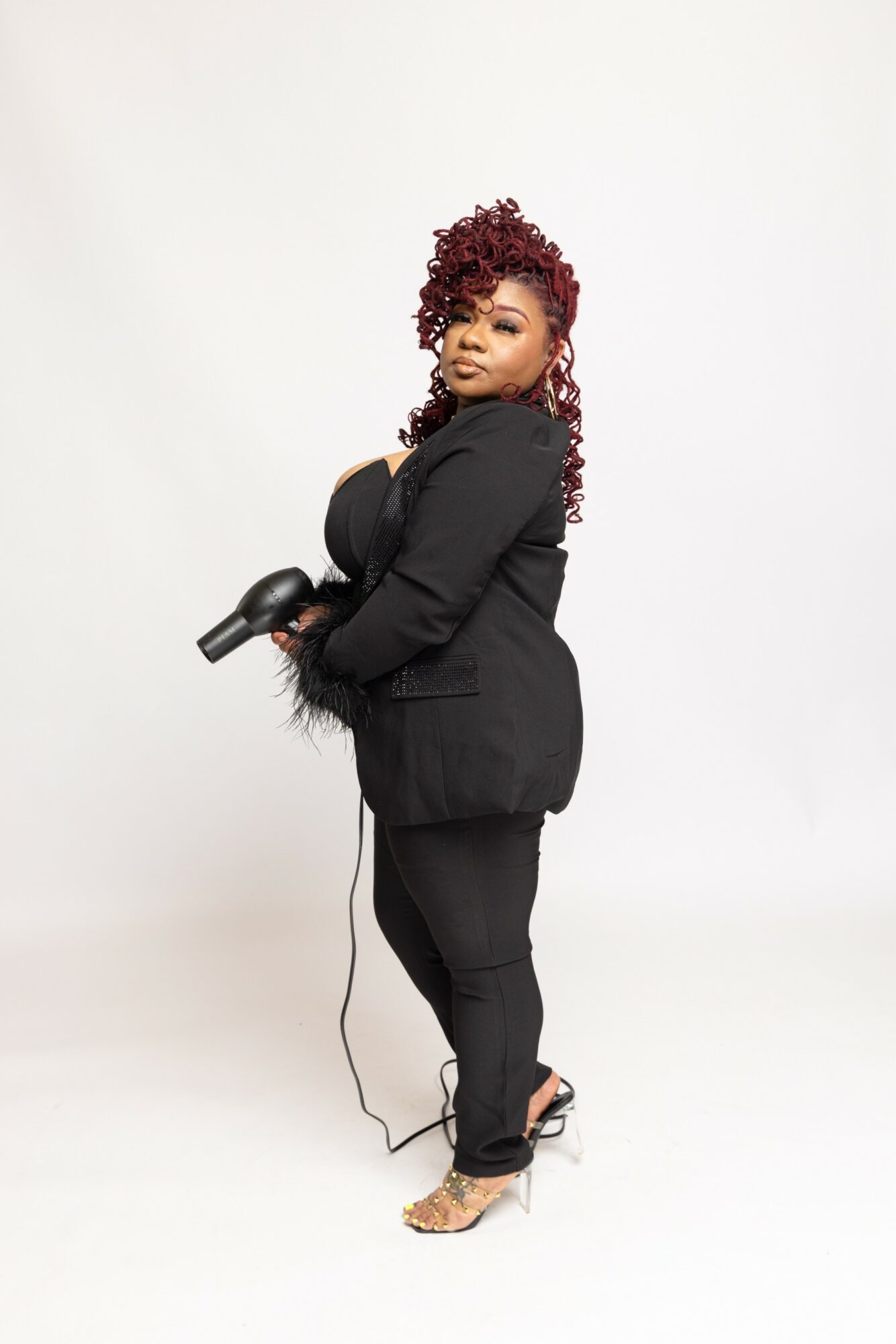 Woman with curly red hair in black blazer and pants holding a hairdryer, standing against a white background.