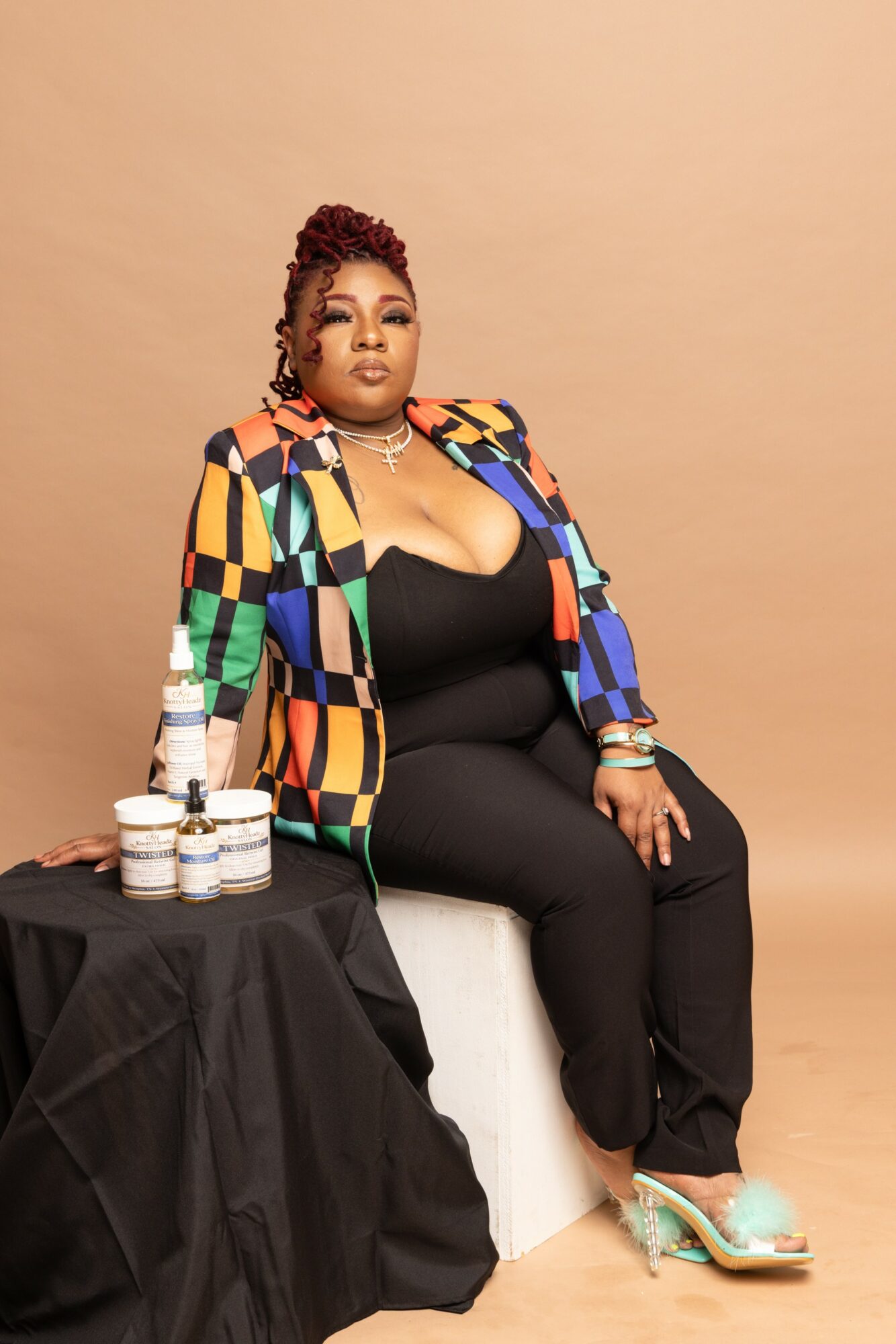 Woman sitting on a white block with skincare products on a black table beside her, wearing a colorful blazer and black outfit.