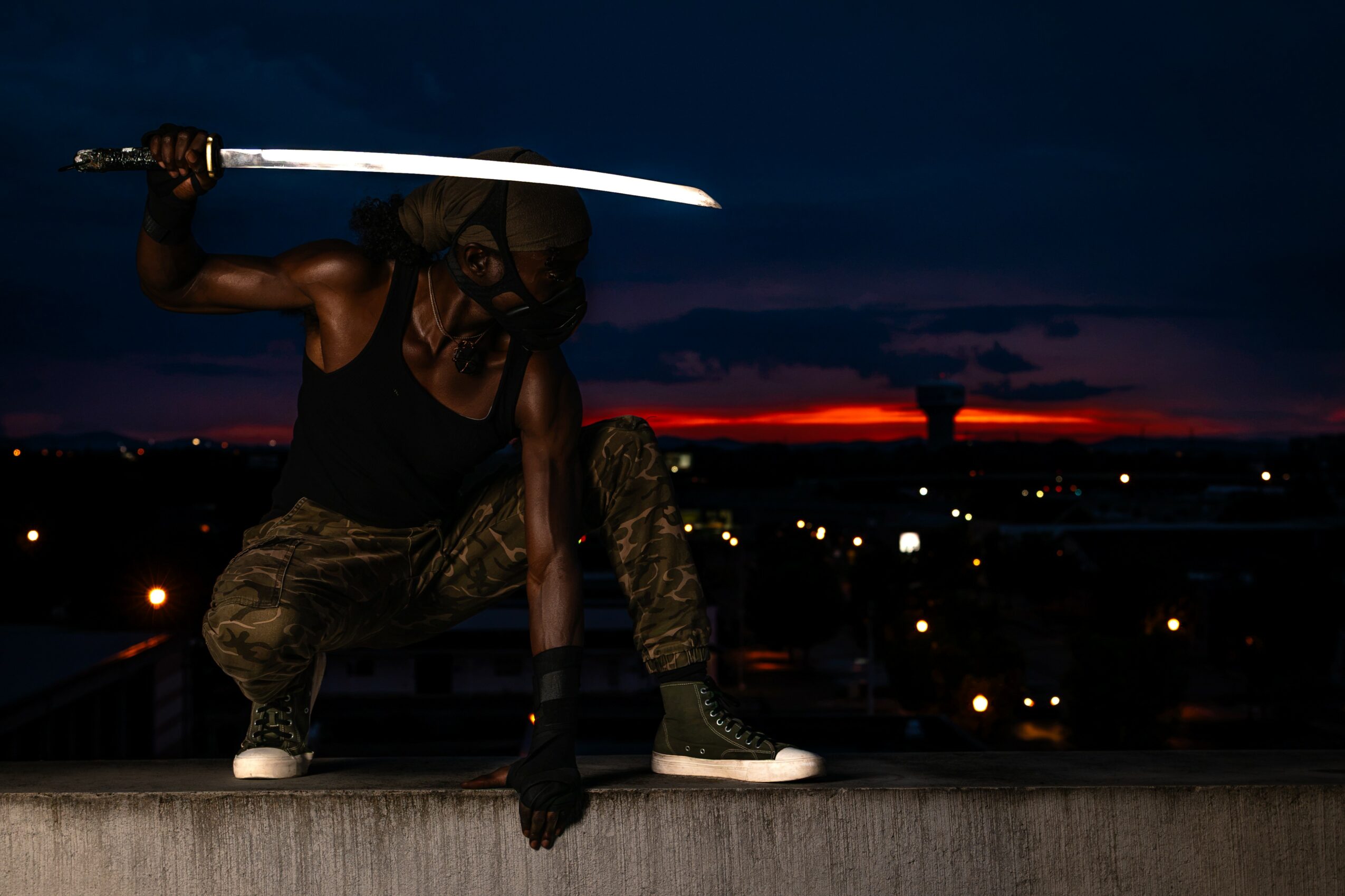 Person crouching on a rooftop holding a sword, with a cityscape and sunset in the background.