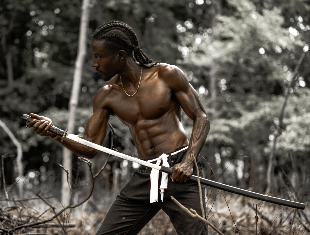 Muscular man with long braided hair holding a sword in a forest setting, looking to the side.