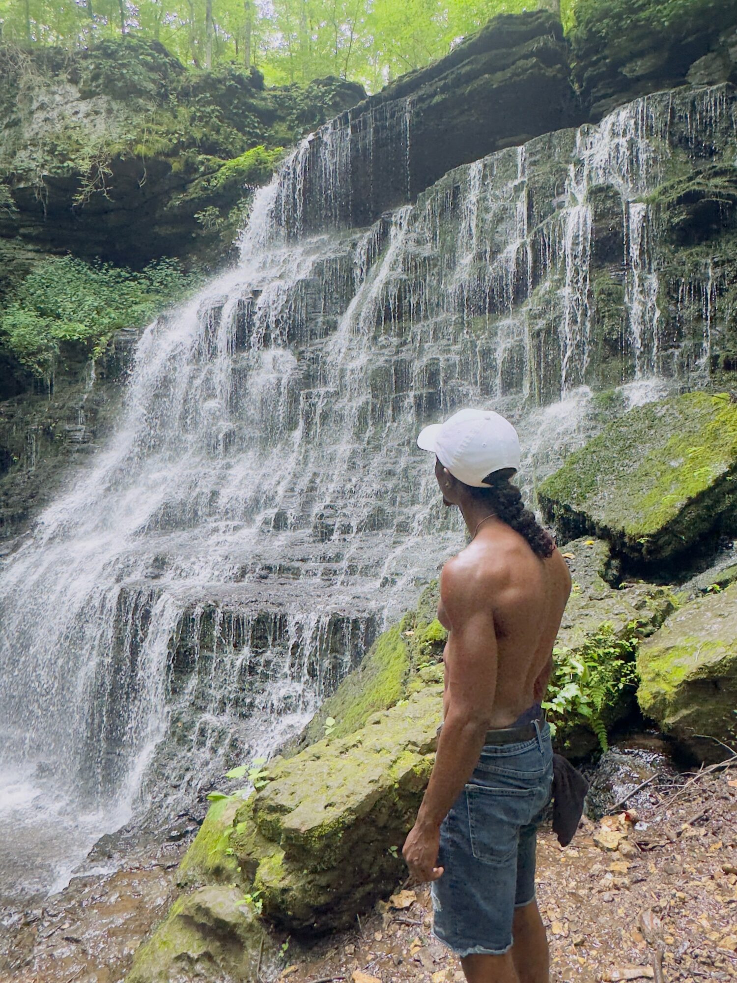 A shirtless man wearing a white cap and denim shorts stands near a waterfall surrounded by rocks and greenery.