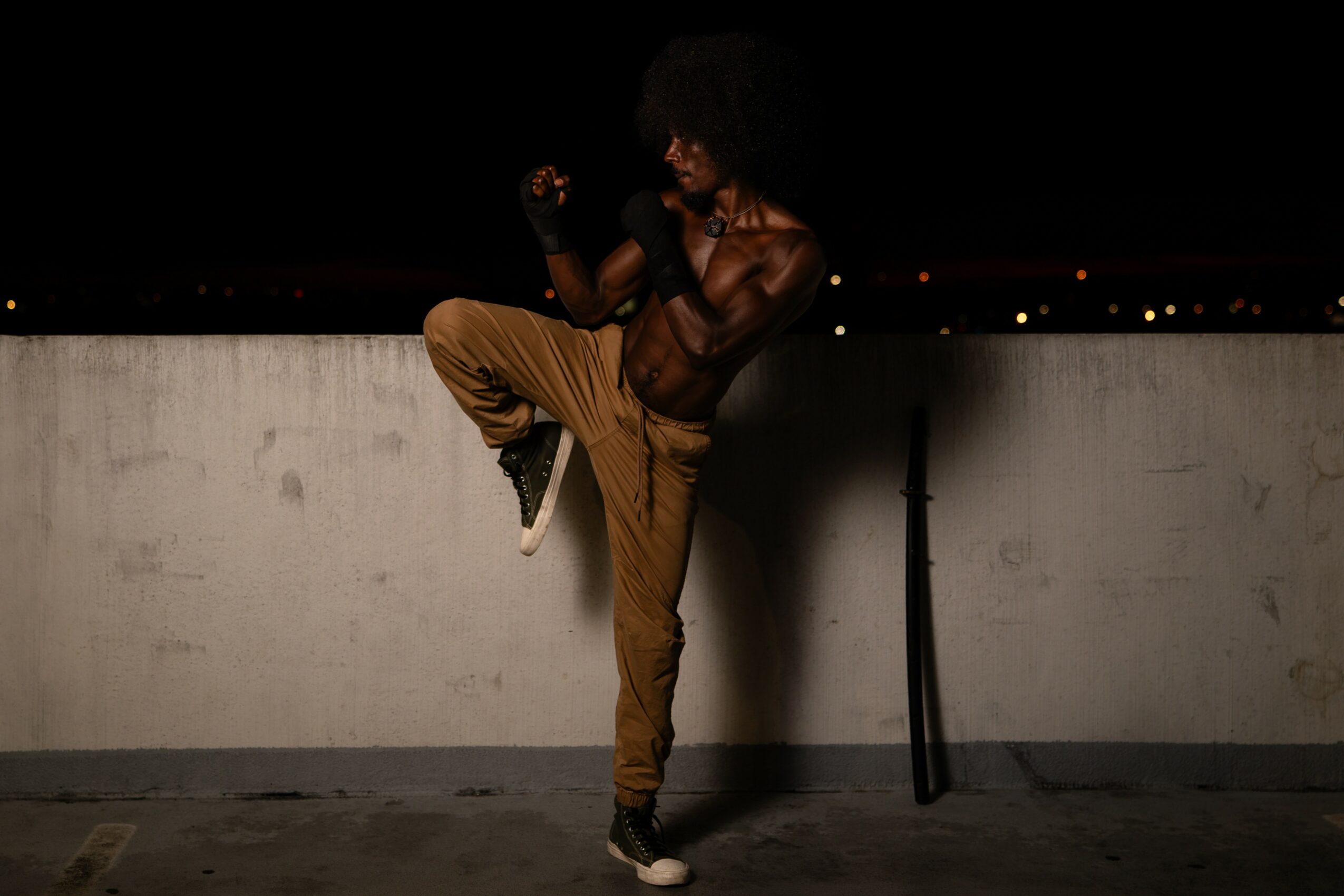 Person with curly hair poses on a rooftop at night, with city lights in the background, wearing a sleeveless top and pants.